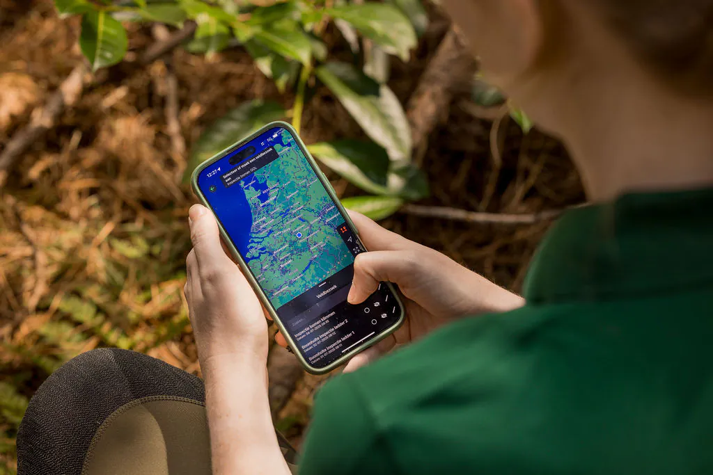 Ecoloog met VeldApp op de telefoon, kaart met plangebied in beeld, gehurkt in de bosrand